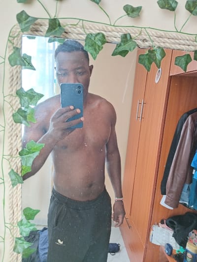 A man takes a selfie in a mirror. He is shirtless, wearing black athletic shorts. The mirror has a rope-like frame decorated with artificial ivy. Behind him is a wooden cabinet or dresser, with clothes piled on top. A water bottle or mug is also visible. The man is holding a blue smartphone.