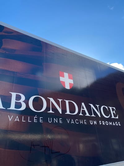 The image depicts a large outdoor banner or sign, partially visible against a bright blue sky with sparse white clouds. The banner features dark brown background imagery, resembling stacked wooden planks or blocks. Prominently displayed in white capital letters is the word "ABONDANCE". Below it, in smaller white capital letters, is the French slogan "UNE VALLÉE UNE VACHE UN FROMAGE" (A Valley A Cow A Cheese). To the right of "ABONDANCE", a red shield logo with a white cross is visible. Faint, dark graffiti-like markings are present on the lower left section of the banner, partially obscuring the background. The photograph is taken from a low angle, looking upwards at the sign and the sky. This banner is located in the city of Abondance, France, promoting the local region and its famous cheese.
