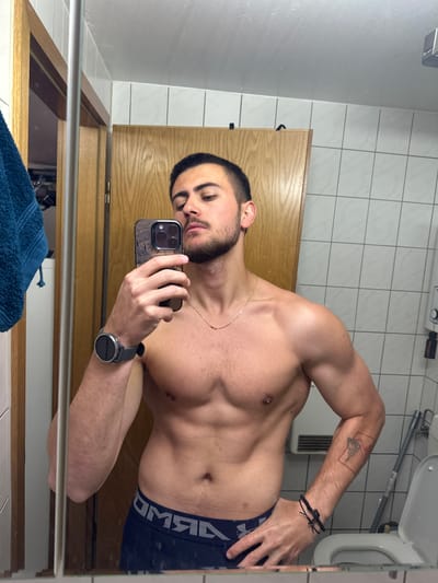 A shirtless man takes a mirror selfie in a bathroom. He is wearing dark-colored underwear with white lettering, a gold chain, and a smartwatch. He has dark hair, a beard, and a tattoo on his left forearm. The bathroom features white tile walls, a wooden door, a white toilet, a towel rack, and a radiator. The city of Essen, Germany, cannot be determined from this image.