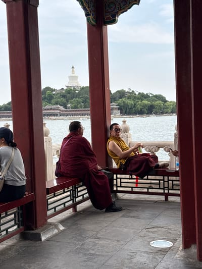 The image shows a group of people in a covered walkway with a view of a body of water and a white pagoda in the distance. The walkway has red painted wooden beams and decorative trim.
In the foreground, a person in a gray shirt and dark pants is hunched over, appearing to be praying or meditating. Further back, two people dressed in maroon robes, likely monks, are visible. One monk is lying down and looking at a phone, while the other is seated and looking out at the view. The monk who is lying down appears to be wearing glasses and has a relaxed posture. The seated monk is facing away from the camera, but their maroon robe suggests they are also a monk.
The background features a wide body of water, possibly a lake, with trees and traditional Chinese architecture along its edge. A prominent white pagoda stands on a hill covered in trees, a likely landmark. The sky is overcast, suggesting a cloudy day. The overall atmosphere seems peaceful and contemplative, with the individuals enjoying the scenery. The location appears to be a temple or a scenic spot in Xicheng District, China. There is no visible text in the image.