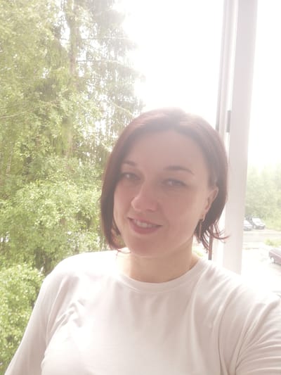 In a selfie taken from the chest up, a woman with shoulder-length brown hair and fair skin is looking towards the camera. She is wearing a white t-shirt and a small hoop earring with a dangling charm in her left ear. The background is bright and appears to be an outdoor scene seen through a window or glass door. To the left, out-of-focus green foliage from trees is visible. To the right, a bright, overcast sky dominates the background, with a sliver of a building or wall on the far right edge. Further in the background, two parked cars are visible, one light-colored and one darker, in what appears to be a residential area. The overall lighting suggests it is daytime, possibly a cloudy or hazy day. This scene is consistent with a residential window view in the city of Tosno, Russia, as indicated by the presence of trees, parked cars, and a typical residential building structure visible in the background.