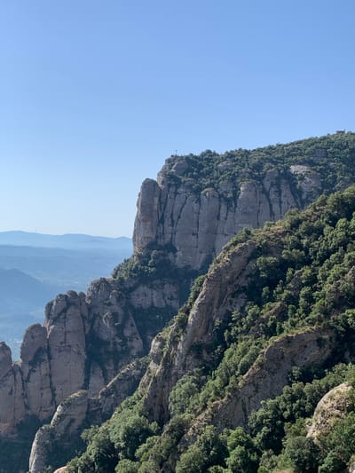 This image shows a panoramic view of a mountainous landscape with rocky cliffs and dense vegetation. In the foreground, a steep, jagged cliff face dominates the right side of the frame, covered in lush green trees. To the left, another series of rocky formations descend into the distance. In the far background, layers of mountains fade into a hazy blue, with a clear sky above. The image appears to be taken from a high vantage point, possibly a hiking trail or viewpoint. It is likely the Montserrat mountain range, located near the town of Collbató, Spain, which is known for its unique rock formations and monastic history. There are no people or animals visible in the frame, and the scene is static and undisturbed.