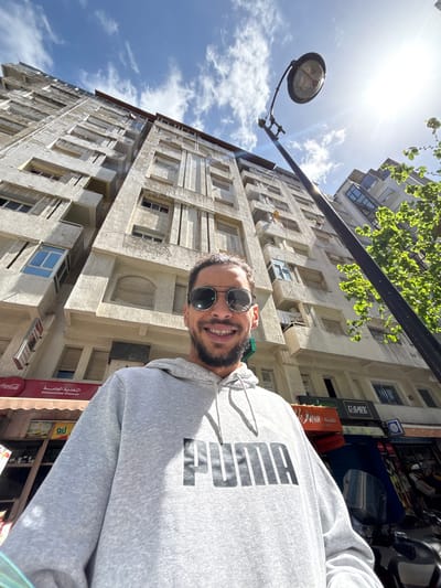 The image presents a low-angle selfie of a smiling adult male in Tanger, Morocco, taken on a bright, sunny day. The man, with short dark hair, stubble, and reflective aviator sunglasses, wears a light grey hooded sweatshirt emblazoned with "PUMA" in black capital letters. He is positioned in the foreground, looking directly at the camera.
Behind the man, a tall, multi-story building with a weathered, light-colored facade rises vertically, dominating the background against a blue sky with scattered white clouds. The building features numerous windows, some with blue glass, and balconies. At its base, visible street-level storefronts include a red Coca-Cola sign, a red awning with "التغذية العامة" (Alimentation Générale - General Food/Store) in Arabic, and a green-and-orange sign partially displaying "U-SAVE." A black street lamp with a circular luminaire extends into the frame from the right, and a tree with vibrant green foliage is visible on the far right. The sun is prominent in the upper right, casting a strong glare. Part of a dark-colored motorcycle or scooter is visible on the far right, near the bottom.