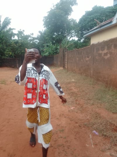 A person holds a smartphone in their left hand while walking on a dirt path. They are wearing a white jacket with red and black detailing, patterned shorts, and one sandal. The path is surrounded by a wall to the right and greenery on the left. Trees and a portion of a building are visible in the background.