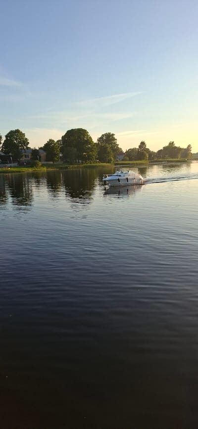 A white motorboat travels across a calm body of water, leaving a wake behind it. The boat is white with black trim and has a covered cabin area. The water is dark and reflects the sky and the trees along the shoreline. The shoreline is lined with lush green trees and some houses. The sky is a clear blue with a few wispy clouds. The scene appears to be set during daylight hours, possibly late afternoon or early evening due to the warm glow of the setting sun on the horizon. The location is identified as Pärnu linn, Estonia.