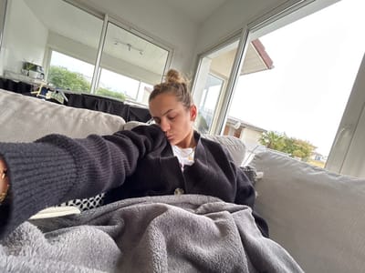 A female individual is depicted resting indoors on a light-colored sofa in Veauchette, France. Her eyes are closed, and her light brown hair is tied in a bun. She wears a dark, ribbed, long-sleeved cardigan over a white inner garment, with a golden chain bracelet visible on her left wrist. A grey, soft-textured blanket covers her lower body and lap. The interior features white walls and a large, white-framed sliding glass door or window in the background. Through the window, an exterior view reveals residential buildings with tiled roofs, green foliage including a palm tree, and an overcast sky. Partially visible in the far left background are several blurred objects on a surface, including a dark rectangular item.
