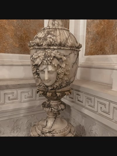 The image shows a close-up of a marble urn adorned with a carved face and grapes. The urn has a classical design and is placed on a pedestal. The background consists of textured walls and architectural details, including a meander pattern. The lighting is soft, highlighting the intricate details of the sculpture. The scene suggests an indoor setting, possibly a museum or an ornate interior space in Saint-Août, France. There are no people or animals visible in the image.