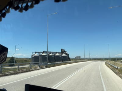 The image shows a view from inside a vehicle, looking out at a road and an industrial facility. The road is paved and has white lane markings. There are several tall, cylindrical silos with conical roofs, characteristic of grain storage facilities, located to the left of the road. These silos are dark in color and appear to be made of metal. A metal guardrail runs along the side of the road. The environment is open and appears to be rural, with dry vegetation and some scattered trees. The sky is clear and blue. Several streetlights are visible along the road. In the foreground, the top of the vehicle's dashboard and a rearview mirror are partially visible. The overall scene suggests a sunny day in an industrial or agricultural area, possibly on the outskirts of a town or city. The location is identified as Unknown, Greece.