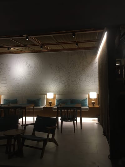 This image shows a dimly lit interior scene, possibly a restaurant or lounge, in Ban Tai Subdistrict, Thailand. The foreground features a dark wooden chair with a woven seat and back, positioned next to a small round wooden side table. Further back, two rectangular tables are visible, each with a woven chair. Along the back wall, a long, built-in bench with light-colored cushions and teal throw pillows is illuminated by a warm lamp on the right. Above the bench is a large mural depicting figures in traditional clothing and a circular motif with a rabbit. The ceiling features a textured material within a wooden grid, with recessed spotlights. The overall ambiance is serene and dimly lit, suggesting a quiet evening setting.