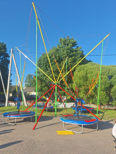 An outdoor scene captured under a clear blue sky features a bungee trampoline setup. The main structure is a colorful framework of red, yellow, and green poles and ropes, from which several bungee cords are suspended. Two trampolines are positioned on the ground beneath the bungee apparatus. The trampoline in the foreground is blue with red and green accents, equipped with a blue harness. Another trampoline, similar in design, is visible further back. In the background, there are trees, houses, cars, and a white picket fence. A person can be seen sitting on the rear trampoline, and a portion of another person is visible on the right side of the frame, facing away from the camera. The ground is covered with gravel and asphalt. The location is identified as Цімкавіцкі сельскі Савет, Belarus.