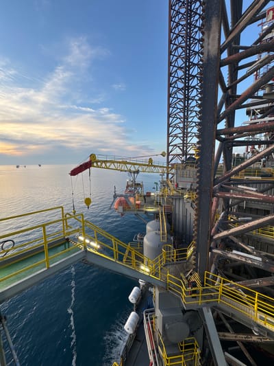 This is a wide-angle, eye-level shot of an offshore oil rig at sunrise. The rig is constructed from dark, weathered metal and features numerous platforms, stairs, and large structural beams. A yellow crane extends from the rig into the frame, with a red boom and a yellow hook. Yellow handrails and lights line the walkways and stairs. In the background, the vast ocean stretches to the horizon, with a few other distant oil rigs visible. The sky is a gradient of blue and orange, with wispy clouds. The water is calm with gentle ripples. The scene is devoid of any visible people or animals. The location is an offshore oil rig, not in a city in Italy.