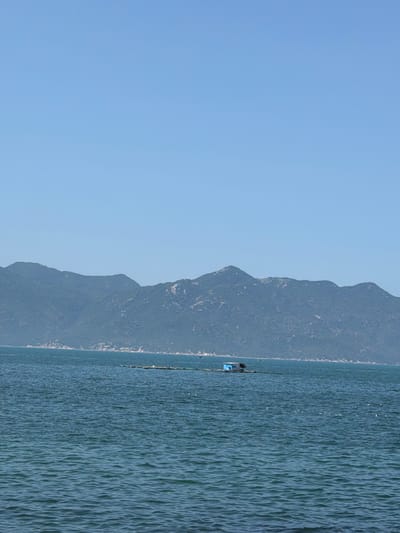 The image displays a maritime scene in Vĩnh Lương, Vietnam, under a clear, light blue sky. A continuous range of dark, heavily vegetated mountains forms the distant background, their lower slopes meeting a discernible rocky coastline. The expansive foreground and mid-ground are dominated by deep blue-green water exhibiting a rippled surface texture. In the mid-ground, a man-made floating structure is visible. This structure consists of a small, light blue, shed-like building with a dark roof, situated upon a dark, elongated platform or a series of interconnected rafts. This entire assembly extends horizontally across the water, consistent with marine aquaculture infrastructure. No individuals are visible within the frame. Scattered small white specks are present on the water's surface.