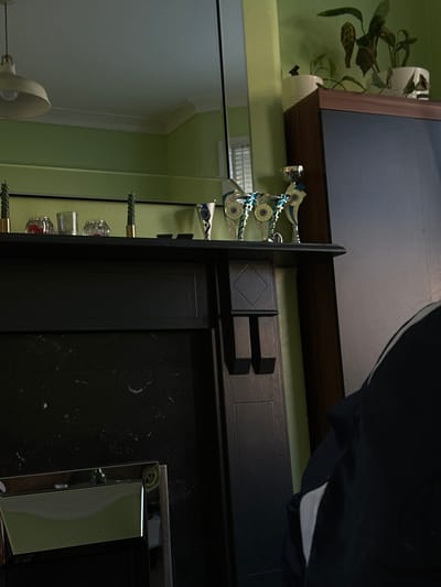 The scene depicts an interior residential space in Dorchester, United Kingdom, focused on a fireplace mantelpiece. The wall behind the mantel is painted a light green. The black mantelpiece features decorative items including two dark green twisted taper candles in brass holders, three clear glass votive holders (two containing small red spherical objects, one empty), and at least four trophies. The trophies consist of one silver-colored cup, one silver-colored trophy with a blue accent, and three glass or acrylic trophies with blue/teal and gold accents, featuring an eye-like design. Above the mantel, a large mirror reflects a cream-colored pendant light fixture, a white ceiling with coving, and a window with white louvered blinds. To the right of the fireplace, a dark brown cabinet with a dark door panel stands, topped by two potted plants in white containers; one plant exhibits brown leaves. In the lower right foreground, the right shoulder and arm of a person, wearing a dark garment with a white stripe, are partially visible and out of focus.