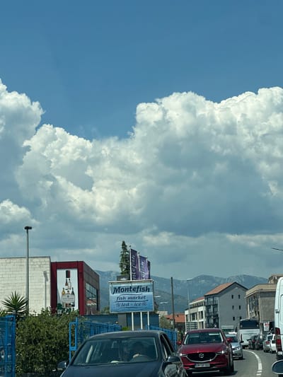 The image captures a congested street scene in Mrčevac, Montenegro, under a sky filled with dramatic clouds. In the foreground, traffic is stopped on a road. Several cars are visible, including a dark colored sedan in the left foreground and a prominent red SUV with license plate "HN BF 068" in the center. Behind the red SUV, other vehicles, including a white van and a bus, are also part of the traffic jam.
On the right side of the road, a white van is parked. The background features buildings, including a white brick building on the left and a building with a red facade displaying an advertisement for wine bottles. A sign for "Montefish fish market" is also visible. In the distance, a mountain range forms the horizon. The overall impression is a typical day in a coastal town, with a busy road, commercial signs, and a striking natural backdrop.