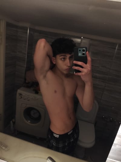 A young, fair-skinned male poses shirtless in a bathroom mirror selfie. He is wearing checkered boxer shorts. His hair is dark and curly. He is holding up a black smartphone, obscuring part of his face. A washing machine and a toilet are visible in the background. The bathroom features gray marbled tiles. The lighting is slightly dim.