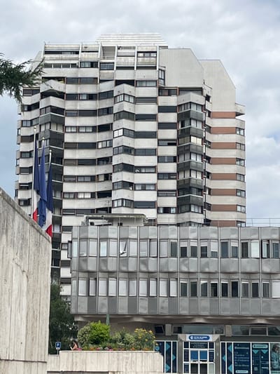 This is a frontal, eye-level shot of a multi-story building in Créteil, France. The building is composed of two distinct sections. The upper section is a tall, white, brutalist-style tower with a stepped facade featuring numerous balconies and windows. It has a white, corrugated material on the upper floors, interspersed with rows of windows. Some balconies have decorative elements. The lower section is a wider, gray, glass-fronted building with rows of rectangular windows, many of which are open. French flags are visible on the left side of the image, in front of the main building. In the foreground, a concrete wall occupies the left portion of the frame. Behind the lower building, there is greenery, including trees and flowers, and a person is visible standing near a parking sign. The sky is overcast. The signage on the lower building indicates it is the "Chambre de Commerce et d'Industrie du Val-de-Marne."