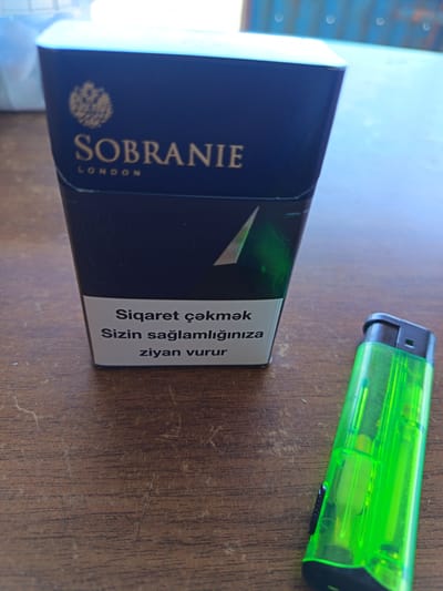 An overhead, close-up shot shows a dark blue cigarette pack with gold lettering and a gold crest on the front, reading "SOBRANIE LONDON". Below this, a white label contains text in what appears to be Azerbaijani, reading "Siqaret çəkmək Sizin sağlamlığınıza ziyan vurur". To the right of the cigarette pack is a translucent green lighter with a black button and a dark grey base. Both items are resting on a dark brown wooden surface with visible grain and scratches. The background is blurred, showing a light blue surface and a darker blue, ribbed object. The scene is lit from above, casting subtle shadows. The location is not discernible from the image content; it is not possible to determine it is in Baku City, Azerbaijan.