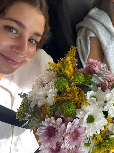 An image captures a close-up perspective inside what appears to be a vehicle. A female subject occupies the left side of the frame, partially visible from the chest upwards. She has medium brown hair, brown eyes, and is smiling towards the camera. She wears a white collared shirt, and a dark seatbelt crosses her chest from her right shoulder to her left hip. To her right, a second individual is partially visible, wearing a light gray hooded sweatshirt. In the foreground and central to the image is a vibrant bouquet of flowers. The bouquet contains white chrysanthemum-like flowers, pink chrysanthemums, bright yellow flowers (possibly solidago), and distinctive round, green-budded flowers (possibly green button chrysanthemums), interspersed with green foliage including eucalyptus leaves. The background is dark and blurred, consistent with the interior of a vehicle. No specific landmarks are visible to confirm the stated location of Aldoar, Foz do Douro e Nevogilde, Portugal.