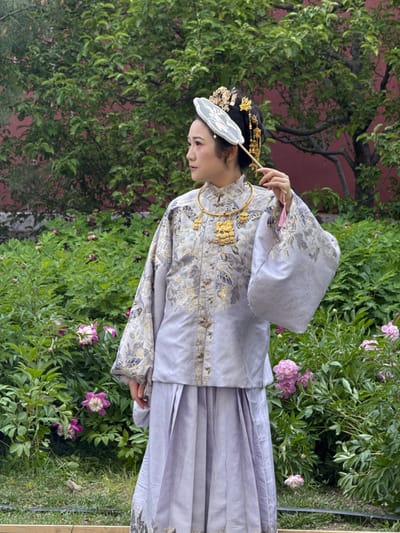 A person in traditional clothing stands among lush greenery in Dongcheng District, China. The individual wears a light purple garment with gold and blue embroidery, along with a golden headpiece and a white fan. The background features a red wall partially obscured by dense green foliage and pink peony flowers. The person is positioned on the edge of a grassy area.