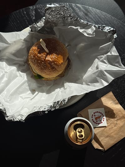 The image presents a burger in a foil wrapper on a white plate, accompanied by a KP Burger logo sticker, a brown paper bag, and an open beverage can. The scene is captured indoors on a black surface. No people are visible. It is not possible to determine the location as Vozdovac Urban Municipality, Serbia from the image alone.