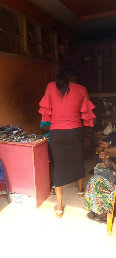 The image depicts an indoor scene within a commercial establishment in Nnewi, Nigeria, likely an electronics repair shop or a vendor of mobile phone components.
In the foreground, a female subject stands with her back to the camera, positioned centrally. She wears a red short-sleeved top with triple ruffles on each sleeve, a black midi skirt, and light-colored slip-on sandals. Her dark hair is pulled back into a ponytail.
To the left of the central subject, a reddish-brown wooden counter holds numerous electronic devices, primarily mobile phones and their disassembled parts, including screens, casings, and circuit boards, arranged in piles. A partially visible person wearing a blue and red top stands behind or next to this counter, obscured by the central subject. A blue plastic chair is visible in the lower-left corner, next to the counter.
To the right of the central subject, a seated individual is partially visible, showing their lower torso and legs. This person wears a colorful patterned skirt (likely Ankara fabric) and dark flip-flops. Their hands are clasped in their lap. A wooden chair leg is visible near their feet.
The environment features reddish-brown or orange-painted walls. In the upper-left background, a wooden shelving unit or window frame contains indistinct items, possibly books or files. The floor is light-colored, with a bright area in the foreground indicating a light source, likely a doorway or window, ahead of the central subject. The rear of the establishment appears darker. No discernible interactions between the subjects are depicted.
