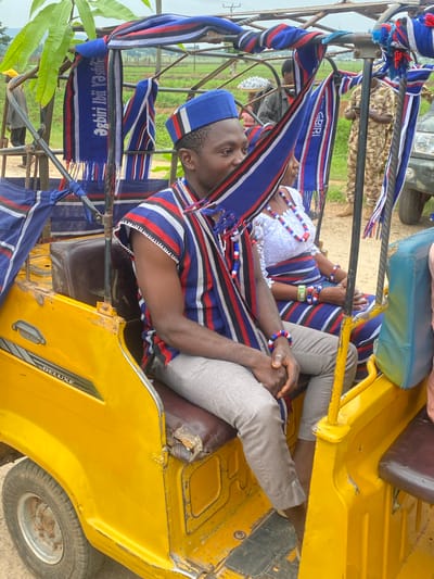 Two individuals are inside a yellow tricycle. The person in the foreground wears a blue hat and a striped shirt of red, white, and blue. They are wearing gray pants, with their hands clasped together. The individual behind them has on white clothing and beaded necklaces and bracelets of white, red, and blue. The tricycle is decorated with striped material of blue, white, and red. The words "agbiri ibi ya state" can be read on one of the decorative pieces, written in reverse. The location could be Maigizo, Nigeria.