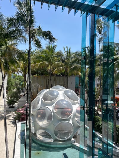 This image captures a bright, sunny day, likely in Miami, with a clear blue sky visible. The scene is dominated by lush palm trees, their fronds casting dappled shadows. A striking, futuristic-looking geodesic dome structure, made of white panels and clear circular windows, is prominently featured. Inside the dome, some interior elements and reflections are visible.
The perspective suggests the photo was taken from an elevated position, looking down through a modern glass canopy. The glass structure itself is supported by dark metal beams and brackets, creating a layered effect. Reflections in the glass show more palm trees and the blue sky.
Below, a pedestrian walkway with a light-colored tiled surface is visible, lined with shops and trees. A few people are casually walking along the path, indicating a leisurely daytime activity. The architecture and abundant greenery suggest a tropical urban environment, consistent with Miami. The overall impression is one of a vibrant and modern outdoor space, bathed in warm sunlight.
There is a partially visible sign with the text "JOHN" on it, likely part of a storefront. The time of day appears to be midday, given the bright sunlight and clear sky. No specific event is evident, but the scene suggests a typical day in a bustling, attractive location.