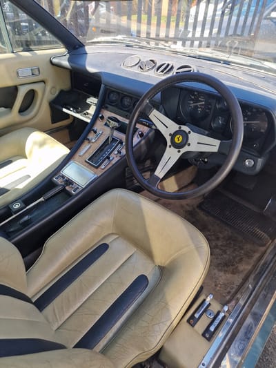 This close-up photograph captures the interior of a classic car, likely a Ferrari given the emblem on the steering wheel, taken from an elevated perspective looking down into the driver's side. The scene is set outdoors in South Tyneside, United Kingdom, under bright, sunny daylight.
The primary subjects are the car's dashboard, steering wheel, seats, and central console. The steering wheel, featuring a black rim and silver spokes, prominently displays the yellow Ferrari logo with a prancing horse. Behind it, the dashboard houses various analog gauges, including a speedometer showing a reading of "52452" miles and marked with "MPH" up to "180," and a tachometer marked "x 1000" up to "80." A switch labeled "LIGHTS" is visible below the steering wheel.
The seats are upholstered in a light cream or beige leather with distinctive dark blue vertical stripes, showing some creasing and wear consistent with age. The central console, partially covered in a wood-grain trim, features a chrome-handled automatic gear selector with positions "P R N D 2 1" clearly visible. Adjacent to the passenger seat, two pull handles are present, one labeled "BENZINA" (Italian for fuel/gasoline) and another, likely for the hood or trunk release, partially visible with "APRI" (Italian for open). The interior appears well-maintained for its age. No people are visible in the image, suggesting the car is parked.