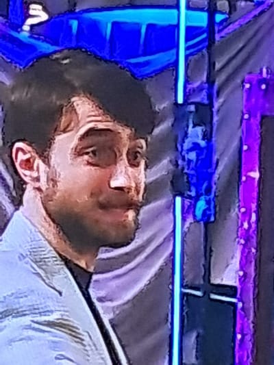 A medium shot, taken from a slightly low angle, shows Daniel Radcliffe from the chest up, looking to his right. He has dark hair and is wearing a light blue blazer over a dark shirt. The background is out of focus and appears to be a stage setting with blue and purple lights and a dark, textured curtain. The image is grainy and appears to be a screenshot from a video. There is no indication of the city of Shopokov, Kyrgyzstan in the image or its description.