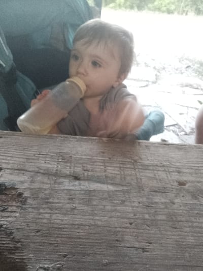 A young child is seated, holding a baby bottle and appearing to drink from it. The child has light brown hair and is wearing a grey shirt. In the background, a large blue backpack is visible. The child is positioned behind a rough wooden table, which occupies the foreground of the image. The setting appears to be outdoors, possibly in a park or natural area, with blurred greenery and light in the background. The lighting is bright and somewhat washed out, suggesting it might be daytime. The image quality is grainy. The specific location in Turgenevskii, Russia, cannot be determined from the visual information provided.