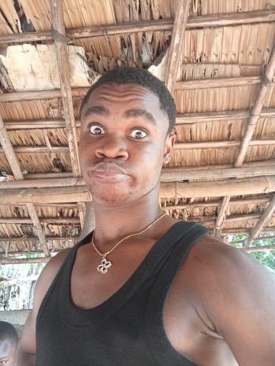 A man with dark skin, short black hair, and expressive eyes is making a playful face. He's wearing a black tank top and a gold necklace with a flower pendant. The background shows a rustic, thatched roof structure made of wooden beams and dried palm leaves. Part of a child's face is visible at the bottom-left. The lighting is natural, giving a warm tone to the scene.