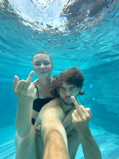 The image depicts an underwater scene in a swimming pool. Two individuals, a woman and a man, are partially submerged. The woman is positioned in front and slightly to the left, wearing a black swimsuit. She is raising her left hand, extending her middle finger. The man is behind her, embracing her, and is also extending his right middle finger. Both individuals have water droplets on their skin and their hair is wet and slightly disheveled due to being underwater. The water is a clear turquoise color, and the pool tiles are visible on the bottom. Bubbles are dispersed throughout the water, and light refracts from the surface, creating a shimmering effect. The overall impression is that of a candid, playful moment captured underwater. The location is specified as Nam Hoa Lư Ward, Vietnam.