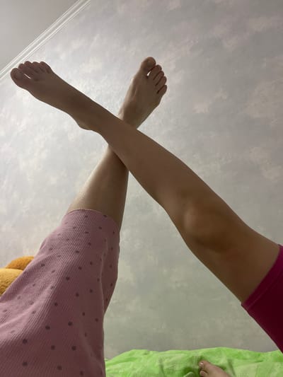 The image shows a close-up of two pairs of legs, one wearing pink polka-dot pants and the other wearing pink shorts, crossing each other. The legs are bare from the knees down, revealing feet with visible toes. One foot is angled upwards, its sole facing the camera, while the other is positioned more horizontally. The background is a wall with a textured, cloud-like pattern. A hint of a green blanket is visible at the bottom of the frame. There is no discernible information indicating the location in the city of Donskoye, Russia.