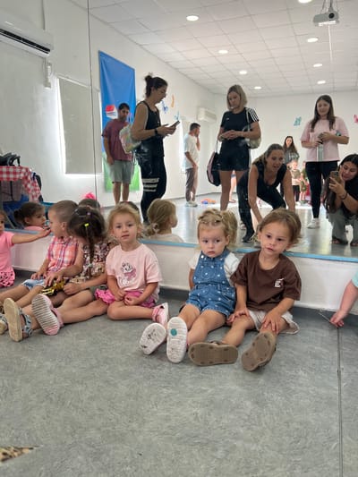In Kiryat Bialik, Israel, a dance studio with a mirrored wall and a projector is the setting for a group of young children and adults. Five young children are seated on the floor, facing the camera. The child in the foreground on the right wears a brown t-shirt and beige shorts, while the child to their left wears a blue denim romper over a white t-shirt. Another child to the left, wearing a pink t-shirt and pink skirt, is looking to the left. Two more children are seated between the first three, one in a pink and white checkered shirt holding a golden toy car, and another in a patterned dress.
Behind the children, several adults are visible, some interacting with their phones. A woman in a black tank top and black shorts is looking at her phone. Another woman in a pink t-shirt is also holding a phone. In the background, two men and another woman are also present. The studio features a white ceiling with lighting and a projector, a white wall with an air conditioning unit, and a large blue banner with a red and white house graphic. The floor is a gray, textured surface.