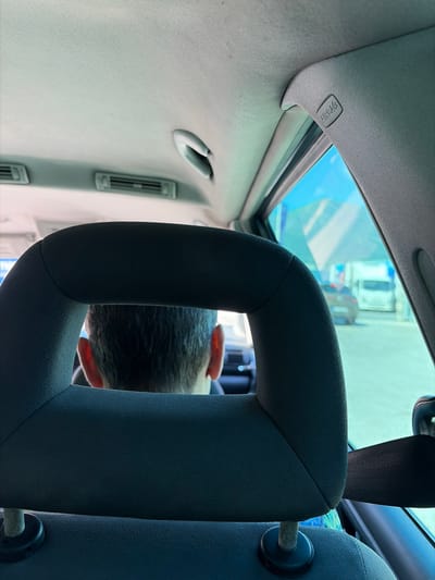 The image captures the interior of a vehicle, looking forward through the rear headrest. A person's head and shoulders are visible in the foreground, facing away from the camera. The headrest is dark grey fabric, and the person has dark hair. Beyond the headrest, a blue sky and several vehicles are visible through the car window, suggesting an outdoor setting. The car's interior includes a grey headliner with air vents. The scene appears to be a snapshot from inside a car, potentially in Dobrota, Montenegro, given the context provided, with a focus on the interior elements and a glimpse of the outside environment.