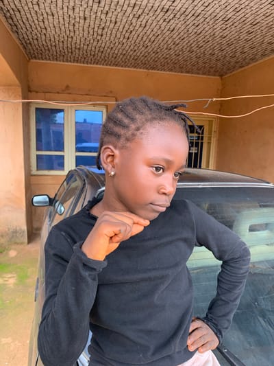 A young Black child is pictured from the shoulders up, looking to their right with a pensive expression. They have braided hair, small hoop earrings, and are wearing a black long-sleeved shirt. Their right hand is raised to their chin, with fingers lightly touching their cheek.
The child is positioned in front of the windshield and side window of a car. The car's exterior is dark and reflective. Behind the car, a building with a rough, textured beige wall is visible, along with two blue-tinted windows and an open doorway. The setting appears to be outdoors, possibly during the day, with a hint of green foliage on the ground in the lower right corner. The overall lighting suggests it's not the brightest part of the day. The context of Barakin Sabawa, Nigeria, places this scene in a specific geographic and cultural setting.