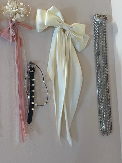 The image displays a collection of fashion accessories laid out on a flat, neutral-colored surface, likely a table or floor. In the top portion of the image, a long, multi-layered bracelet or choker made of small, shimmering rhinestones is positioned horizontally. Below this, a large, cream-colored satin bow is draped, its fabric flowing outwards. Further down, a black choker adorned with small pearls and silver spikes is visible. At the bottom of the image, a delicate floral-shaped hair accessory or embellishment made of pearls and gold wire is partially visible, connected to a pink ribbon bow. Several strands of thin pink and white ribbons are also spread out below the choker and floral accessory. The overall presentation suggests a retail display or a personal collection of decorative items. There is no visible text in the image, and the lighting appears to be even, indicating an indoor setting. The context provided indicates this image was taken in Svilengrad, Bulgaria, but there are no specific visual cues within the image to confirm this location.