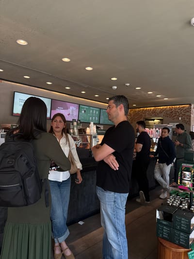 The image captures the interior of a coffee shop, identified as being located in San Agustín, Mexico. The scene features seven individuals engaged in various activities within the establishment. In the foreground, a woman with long dark hair, wearing a dark green long-sleeved top and a matching pleated skirt, is positioned with her back to the viewer, carrying a black backpack monogrammed with "AB." She faces a woman to her right, who has long brown hair, a white collared shirt, cuffed blue jeans, and light brown flat shoes, holding a smartphone. Adjacent to them, a man with short gray hair and a beard, dressed in a black t-shirt and blue jeans, stands with his arms crossed, looking towards the two women.

The background reveals a coffee bar counter with multiple digital display screens showing menus and promotions in Spanish, including "NUEVO Chai Cherry." Various coffee-making equipment and serving items are visible on the counter. Further down the counter, several other male patrons are present: one in a black t-shirt with a tattoo on his left arm, another in a dark blue jacket and light pants, both leaning against the counter, and two partially visible men in the rear, one in a green shirt. The ceiling is light-colored, fitted with numerous recessed circular lights and a smoke detector. A decorative wall panel with an earthy-toned floral or vine pattern is visible in the background. On the right side of the foreground, a wooden retail display stand features mugs, some bearing "Mexico" branding, and boxed coffee products. The floor is dark-colored.