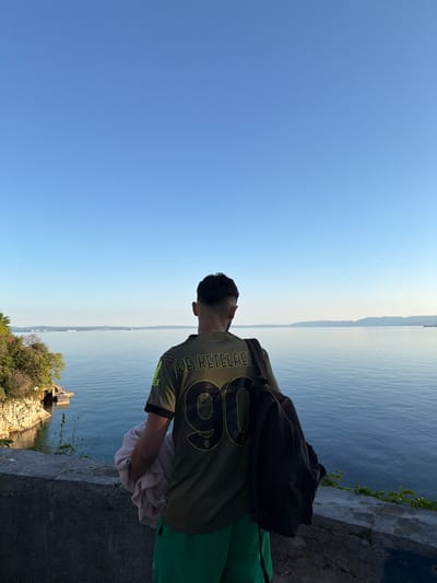 A man wearing a green jersey with the number "96" and the words "DE KETELAERE" stands on a concrete ledge overlooking a body of water, presumably the Adriatic Sea, in the city of Grad Rijeka, Croatia. He has a black backpack slung over his shoulder and holds a white towel in his hands. The sky is clear and blue. In the distance, there are mountains or islands visible across the water. Below the ledge, there is a rocky area with some vegetation. The scene is well-lit, indicating a sunny day.