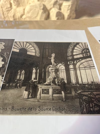 This is a black and white photograph of the Buvette de la Source Cachat in Évian-les-Bains, France. The Buvette is an ornate building with a grand interior featuring large arched windows and intricate architectural details. In the center of the image is a fountain with a statue of a nude figure on top. Several people are present in the scene, dressed in period clothing, including men in hats and suits, and women in long dresses and hats. Some individuals appear to be standing by the fountain, while others are seated at tables in the background, suggesting this is a public space where people gather. The overall atmosphere appears to be one of elegance and social gathering. The caption at the bottom of the photograph reads "- Buvette de la Source Cachat".