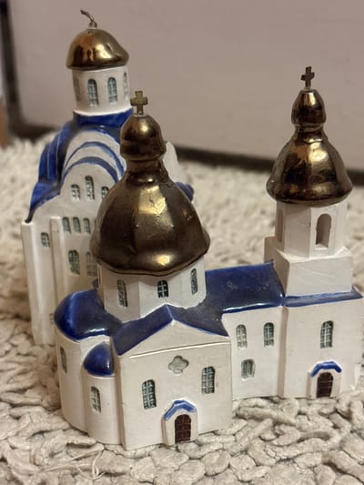 This is a close-up, vertically oriented image of a decorative miniature model of a building, likely a church or cathedral, with distinct architectural features. The model is primarily white with blue accents, and features two prominent golden domes topped with small crosses. The building has multiple levels with numerous small, rectangular windows that appear to be gridded. Blue, curved architectural elements are integrated into the white structure, resembling archways or decorative trim. The model is placed on a textured, off-white surface that resembles a knitted fabric or rug. The lighting appears to be artificial and soft, with no strong shadows, suggesting an indoor setting. There is no discernible text in the image. The overall impression is of a delicate, handcrafted item, possibly a souvenir or decorative piece. The location context of Novaya Usman', Russia, suggests this building might be a representation of a local landmark.