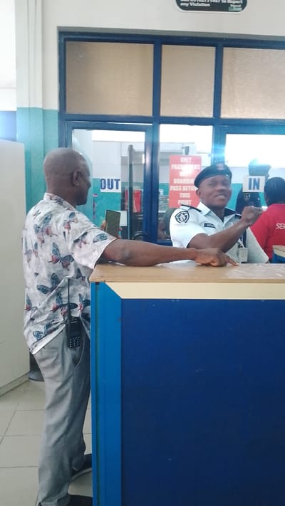 In Orerokpe, Nigeria, a man in a patterned shirt and grey pants is at a counter, holding a mobile phone. Facing him is a uniformed security officer with a badge that reads "AGADE" and "NIGERIAN SECURITY" on his white shirt, and a black beret. The security officer is smiling and gesturing. Behind them, a sign indicates "ONLY PASSENGERS WITH BOARDING PASS BEYOND THIS POINT." Another sign in blue says "IN" and a sign in a similar font says "OUT." The environment appears to be an indoor facility, possibly an airport or transportation hub, with tiled floors and fluorescent lighting. A blue counter separates the two individuals.