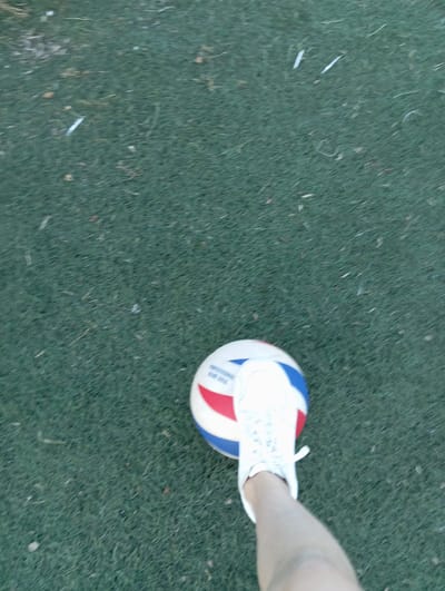 A person's leg and foot, clad in a white sneaker, are positioned over a volleyball. The volleyball is red, white, and blue. The scene is set on an artificial turf field. The image appears to have been taken from a high-angle perspective, looking down at the leg and ball. There is no discernible information about the specific location within Radzymin, Poland, from the visual content of the image itself.