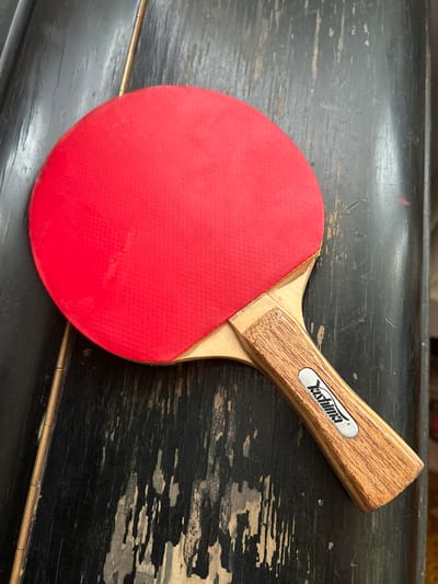 A red ping pong paddle with a wooden handle is placed diagonally on a dark, weathered wooden surface. The paddle's red rubber surface faces upwards, showing a faint textured pattern. The handle is light brown with visible wood grain and features a silver metallic emblem with the brand name "YASHIMA" inscribed in black. The wooden surface appears to be a table or a similar flat object, with noticeable wear, scratches, and peeling paint, revealing lighter wood underneath. The lighting is consistent and casts subtle shadows. The scene is a close-up, top-down view of the paddle and the surface it rests on.