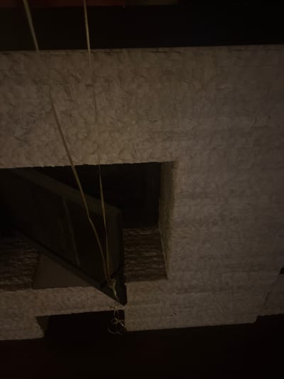 The image shows a close-up view of a textured, light-colored stone wall, possibly a facade or interior finish. A dark rectangular opening is visible in the wall, revealing a dark interior space. Two thin yellow strings or wires hang down from the top of the image, passing through the opening and disappearing into the darkness below. The stone pattern is irregular and appears to be composed of stacked rectangular pieces. The lighting is dim, casting shadows and obscuring details within the opening. There is no discernible information about a specific location in the city of Orekhovka, Russia, or any people, specific objects, or actions beyond the visual elements described.