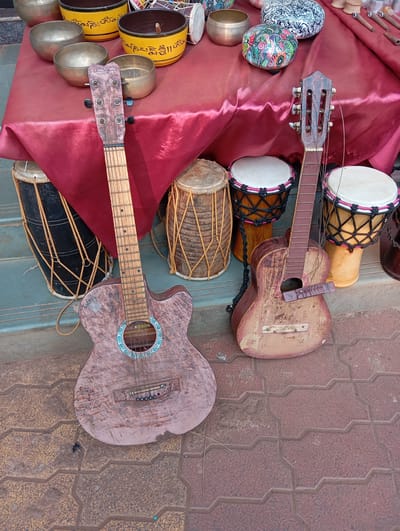 This image captures a vibrant, rustic display of musical instruments and crafts, likely from an outdoor market stall in Arambol, India. The foreground is dominated by two aged acoustic guitars leaning against a low table covered with a deep red tablecloth.

The guitar on the left is a light-colored wooden acoustic model, exhibiting significant wear, scratches, and a distressed finish. It features a decorative turquoise and white rosette around its soundhole. A small white sticker above the soundhole reads "GodSon," and a label visible through the soundhole states "GODSON MUSICAL INDUSTRY." The guitar on the right is a smaller, equally worn acoustic guitar, with a detached bridge and broken strings, suggesting disrepair or a decorative purpose.

Behind the guitars, on a slightly elevated surface or steps, several drums are arranged. These include tall, dark-stained cylindrical drums with string lacing, and multiple djembe-style drums with white drumheads and intricate black and pink lacing patterns around their bases.

The red-draped table holds an assortment of items. Numerous brass singing bowls are visible; some are plain, while others are painted bright yellow with black Tibetan script, most likely "Om Mani Padme Hum." Also on the table are decorative, round, painted containers or bowls – one featuring intricate white and black patterns, and another adorned with colorful, vibrant floral designs. To the far right of the table, a collection of smaller wooden and metal objects, possibly flutes, pipes, or craft tools, are laid out.

The ground surface is a reddish-brown tiled pavement with a distinct zig-zag or interlocking pattern. No people are visible in the frame, and the lighting suggests daytime. The overall scene evokes the atmosphere of a casual market or street vendor's display of local handicrafts and instruments.