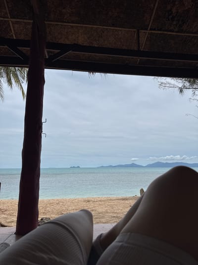 The image depicts a tranquil scene from a beachside shelter in Baan Tai, Thailand. In the foreground, a person's legs are visible, suggesting they are reclining. The shelter's thatched roof and wooden beams are in the upper portion of the frame, casting shadows. A dark, textured pillar stands to the left, partially obscuring the view of the beach and the ocean. The beach itself is sandy with scattered debris. Beyond the beach lies the calm, turquoise ocean, extending to the horizon where a range of islands and mountains are faintly visible under a cloudy sky. The overall atmosphere is one of relaxation and natural beauty.