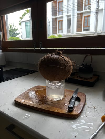 The image depicts a kitchen counter in Chang Phueak, Thailand, with a whole coconut precariously balanced on a glass of coconut water. A wooden cutting board is positioned beneath the glass, with a knife resting on it. The counter is wet, with spills and droplets scattered around, suggesting recent activity involving the coconut. In the background, a window reveals a view of a modern building and some greenery, indicating an urban setting. The overall scene is one of preparation or consumption of fresh coconut water in a domestic environment.