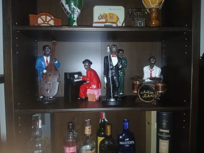 The image shows a dark wooden shelf with various decorative items. The top shelf features a green vase, a tin with an elephant design, an old-fashioned telephone, and a drum. The middle shelf displays a jazz band figurine set. The figurines include a man playing the double bass, another at the piano, a singer with a microphone, and a drummer. They are all depicted as African American men, dressed in suits and formal wear. The bottom shelf holds several bottles of alcohol. One bottle is labeled "MASTER'S GIN," and another is a dark bottle with a gold cap. A tall, dark cylinder with the text "GLENGOYNE" and "CASK STRENGTH 12" is also visible. The overall scene is a collection of decorative objects, suggesting a home or display setting. There are no strong indicators of time of day or weather. The mood is still and contemplative, focusing on the display of these items.
