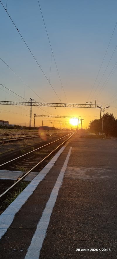 The image depicts a train station platform at sunset in Armavir, Armenia. The sun is low on the horizon, casting a warm, golden glow across the scene and creating lens flare on the train tracks. The platform is made of asphalt with white painted lines indicating the edge. Adjacent to the platform are multiple sets of train tracks, disappearing into the distance. Overhead, a complex network of electrical wires and supporting structures is visible against the clear, pale blue sky. In the background, there are some buildings and trees, as well as distant mountains. The overall atmosphere is serene and suggests the end of a day. The timestamp at the bottom right indicates "20 июня 2026 г. 20:16", which translates to "June 20, 2026, 20:16". There are no people visible in the image.